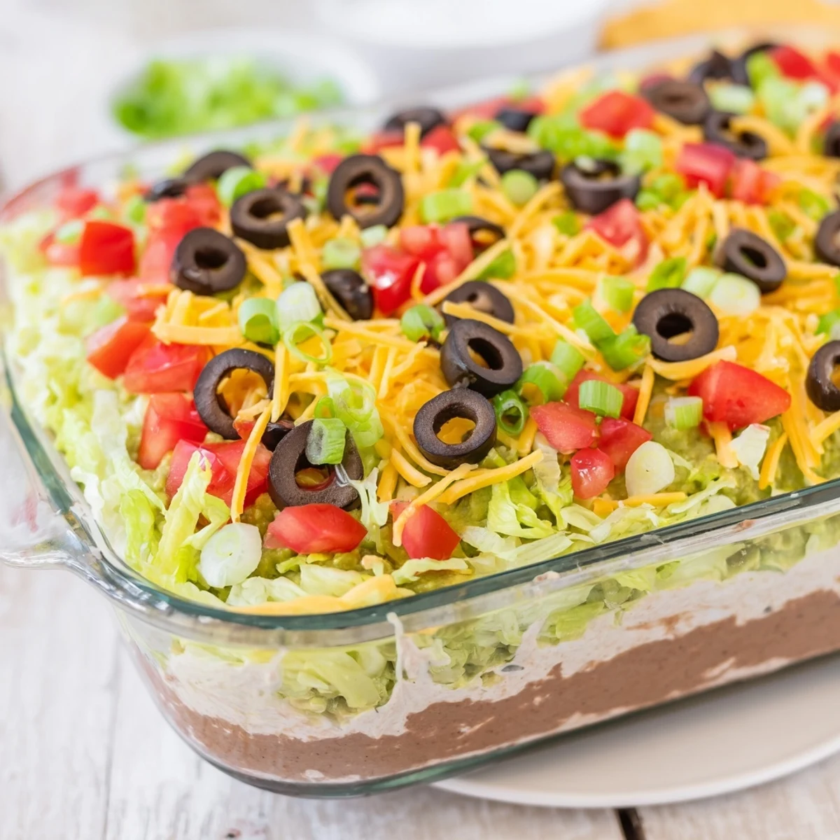 Fresh lettuce, tomatoes, and black olives top the chilled Ultimate Seven Layer Taco Dip for a festive appetizer display.