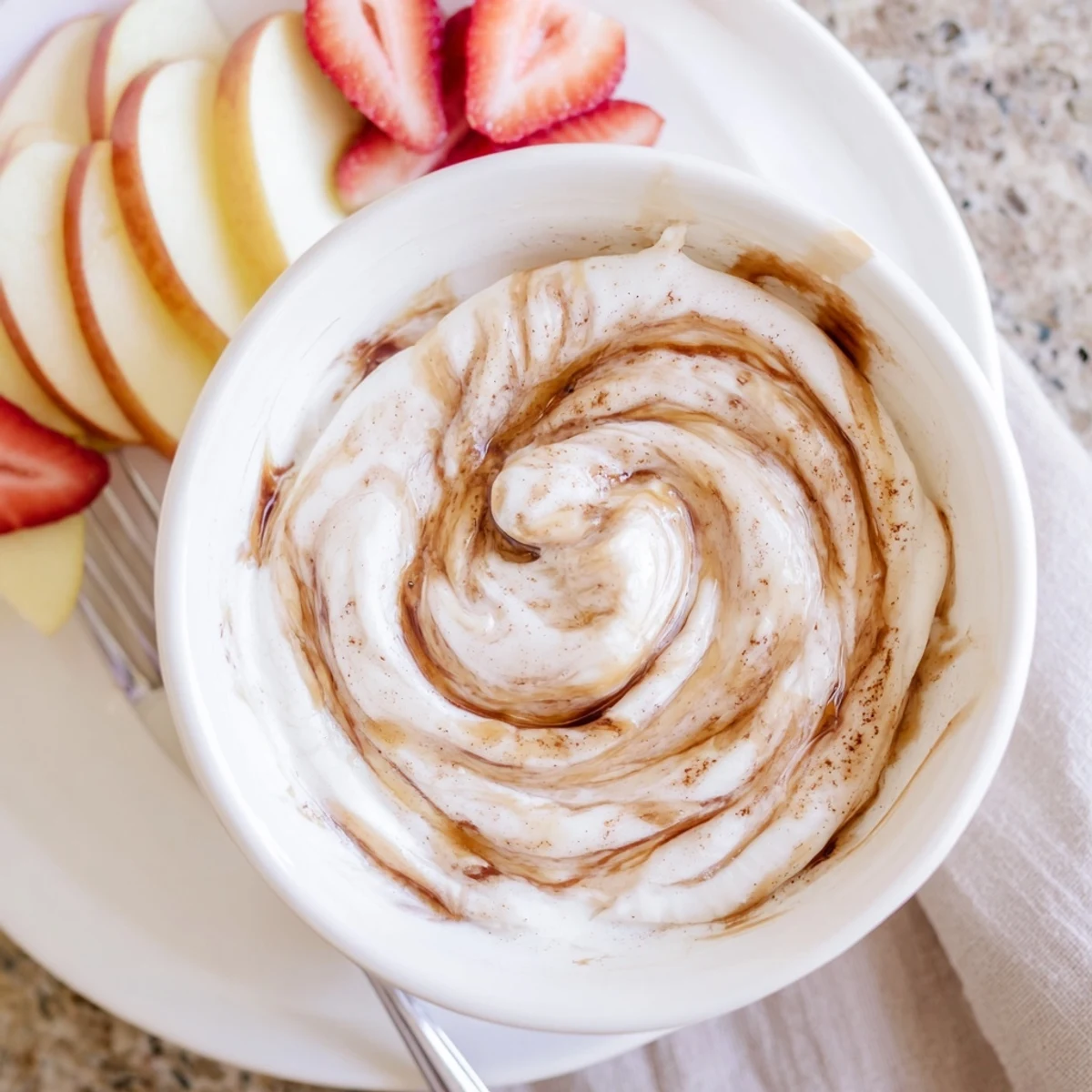Creamy Greek yogurt cinnamon roll dip swirled with cinnamon and served with fresh apple slices and strawberries.