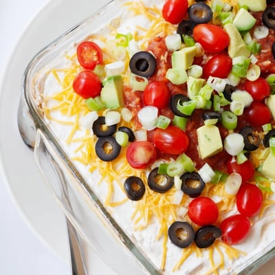 In a 9x13-inch dish, Ultimate Layered Taco Dip showcases refried beans, seasoned sour cream, and fresh avocado, ready for a party platter.