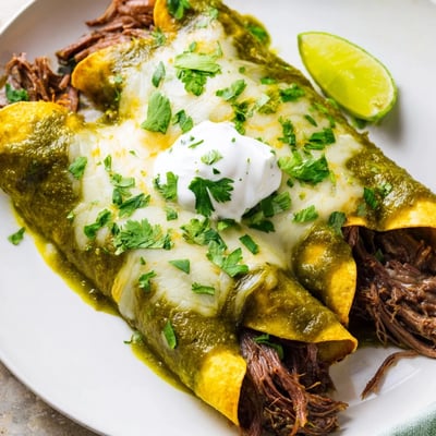 A close-up of Beef Enchiladas with Green Sauce showing melted cheese, cilantro, and lime wedges on a rustic plate.